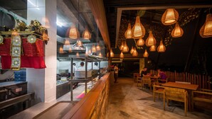 Interior de restaurante com área de bar, mesas de madeira, luminárias pendentes e cozinha visível através de um painel de vidro.