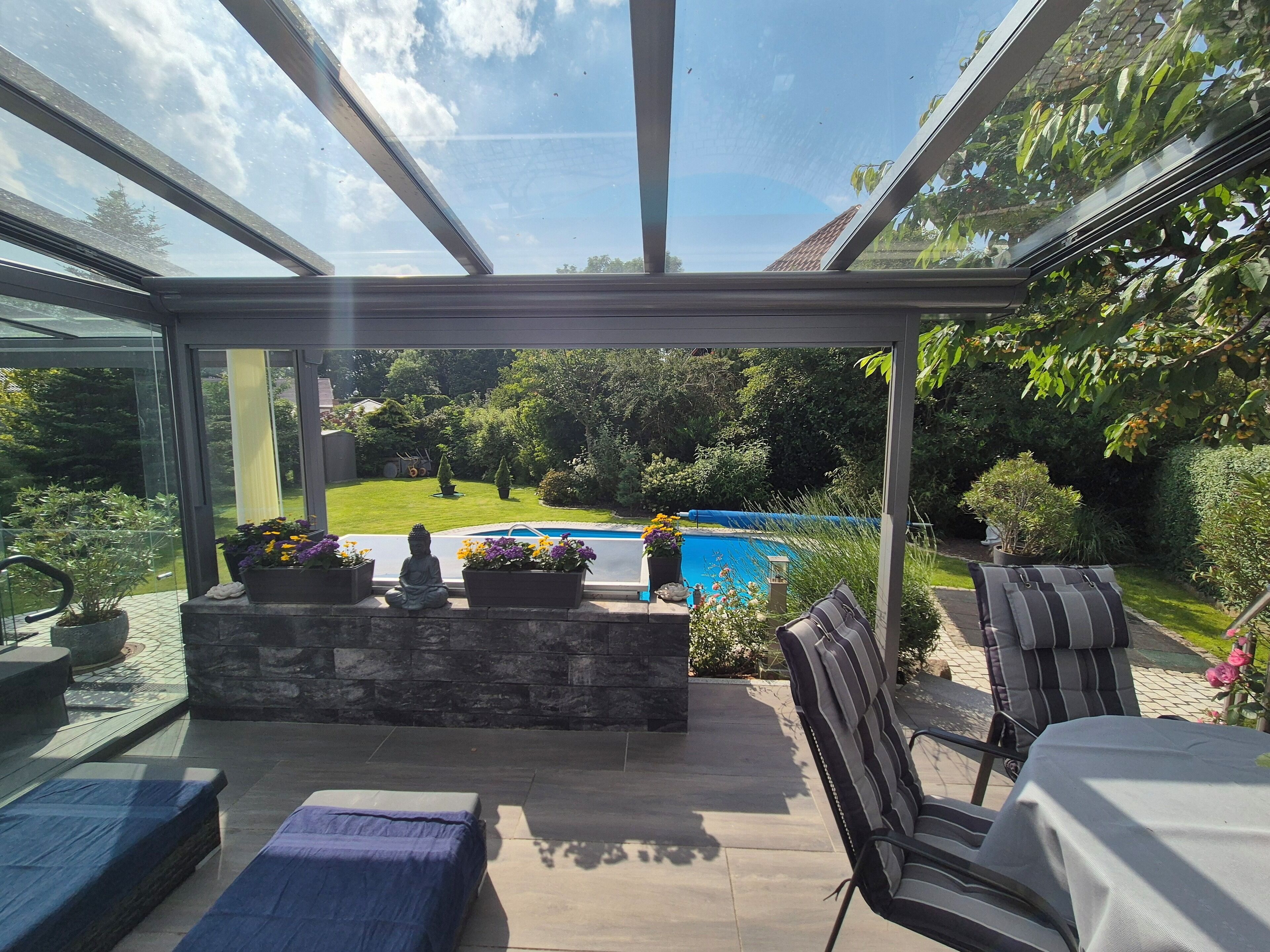 Une véranda fermée par des baies vitrées, offrant une vue sur une piscine, avec un espace salon extérieur et une cheminée en pierre.
