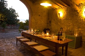 A long dining table set for a meal in a stone-walled room with a view of the outdoors.