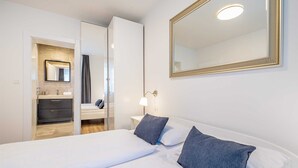 Une chambre d’hôtel avec un lit, une table de chevet, une lampe, un miroir et une salle de bain visible à travers une porte ouverte.