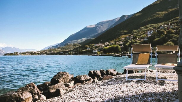 Une vue sur un lac, avec une rive rocheuse, une eau d’un bleu limpide et des montagnes en arrière-plan.