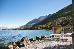 A lakeside view with rocky shore, clear blue water, and mountains in the background. - Renovated rustic house directly on the lake. CIR:023045-LOC-00204 (Malcesine)