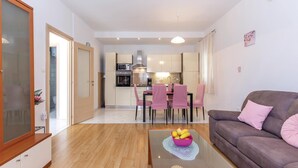 Ein modernes Wohnzimmer mit einem braunen Sofa, einem Glastisch mit Früchten und einem Essbereich mit rosa Stühlen, im Hintergrund befindet sich eine Küche.