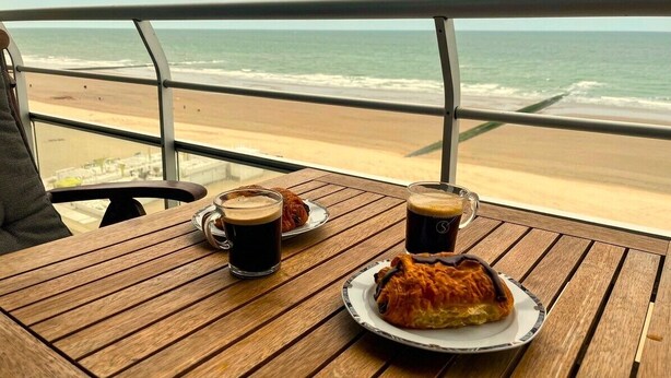 A wooden table with a pastry and two cups of coffee, overlooking a beach.