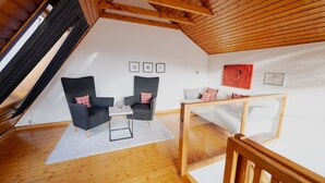 A loft-style room with a sloped ceiling, wooden beams, a white sofa, two armchairs, a small table, and a glass coffee table. - Light-flooded architect house in a quiet location FeWo "Red" (Edertal)