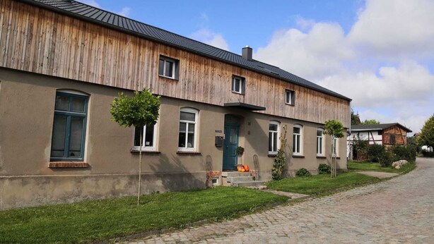 A two-story wooden building with a green door and windows, a cobblestone driveway, and a small tree in front.