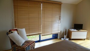 A bedroom with a large bed, a chair with an American flag design, a bedside table with a TV and a mirror, and a window with blinds. - Doëlan: Charming house facing the sea  (Clohars-Carnoët)
