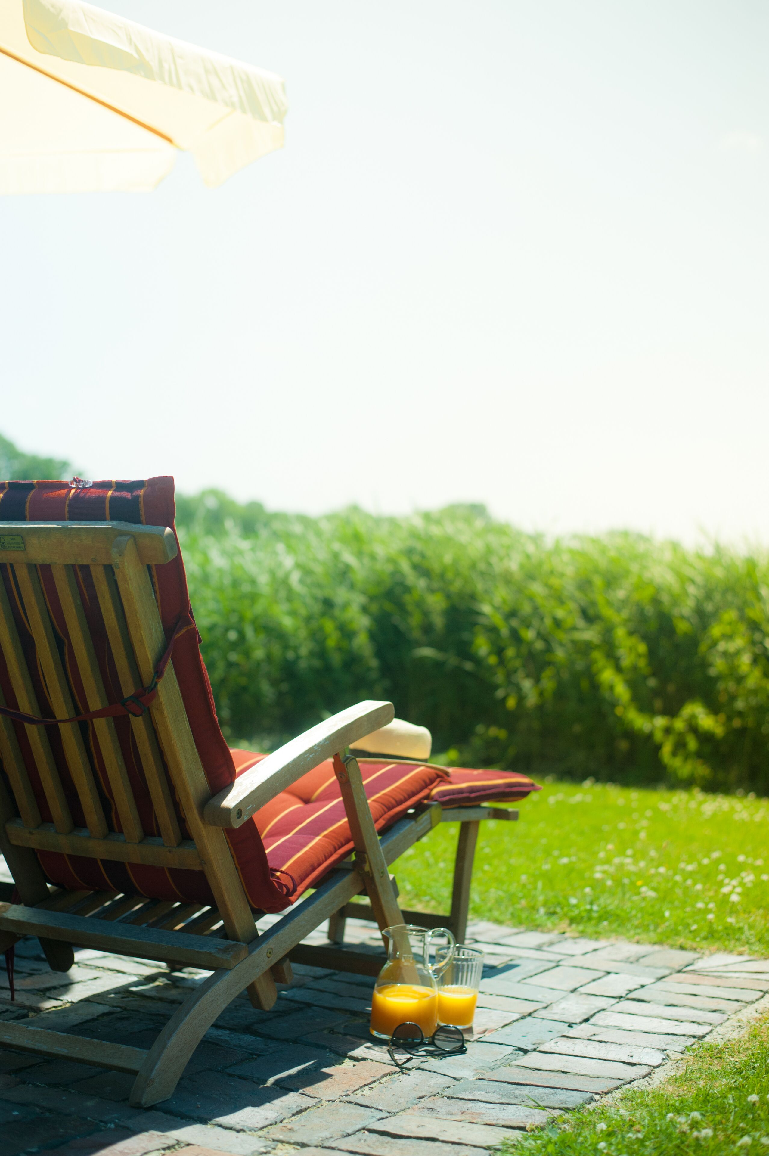 Een houten lounge stoel met gestreepte kussens en een bijpassend voetenbankje, op een terras van bakstenen, met een glas sinaasappelsap en zonnebril ernaast.