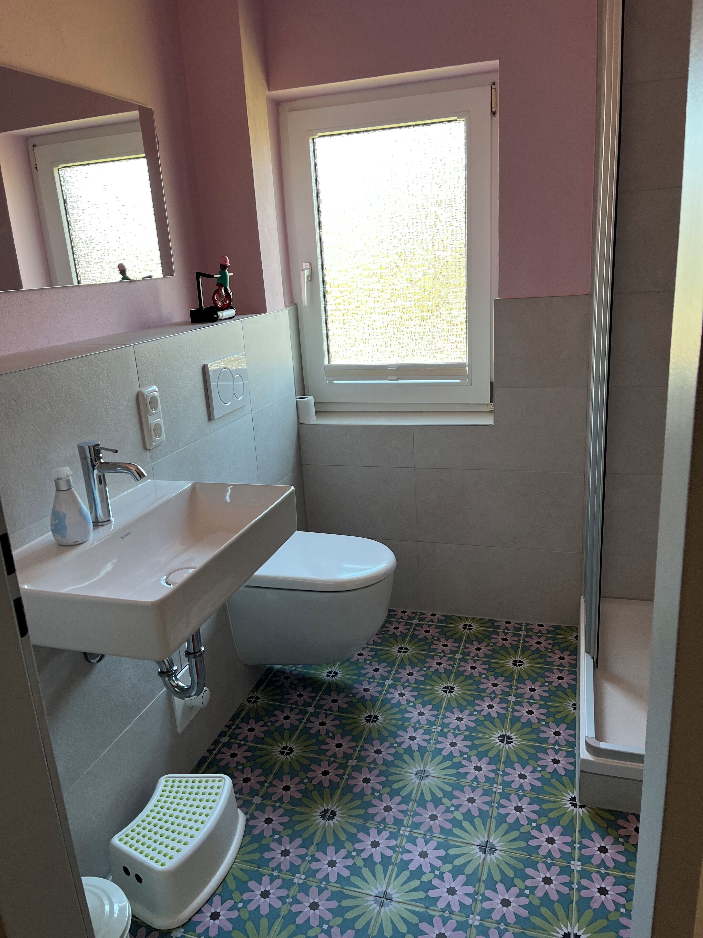 Une salle de bain avec un lavabo, des toilettes et une douche, le tout de couleur blanche. Elle comprend une fenêtre donnant sur l’extérieur, un miroir au-dessus du lavabo et un carrelage au motif décoratif.