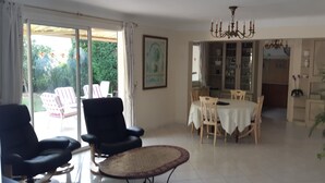 A spacious living area with a dining table, chairs, and a large window with a view of greenery. There are two black armchairs and a woven coffee table. - Fenced garden with 10x4 pool, 85m2 terrace and shaded terrace (Générac)
