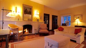 Een gezellige woonkamer met een open haard, een witte bank, een rieten fauteuil en een bijzettafeltje.
