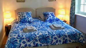 Een tweepersoonsbed met een blauw bloemenbedekt laken, twee bedlampjes, een houten vloer en een raam met gordijnen.