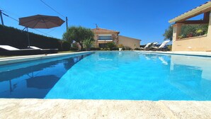 Une piscine entourée de chaises longues et d’un parasol, le tout bordé d’un bâtiment à toit de tuiles et d’un jardin.