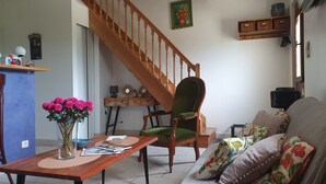 Un salon avec un escalier en bois, un fauteuil recouvert d’un tissu vert, un canapé agrémenté de coussins à motifs floraux, une table basse en bois et un vase contenant des fleurs roses.