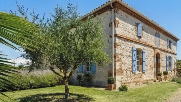 Un bâtiment en pierre avec des volets bleus, un arbre dans le jardin et un ciel dégagé.