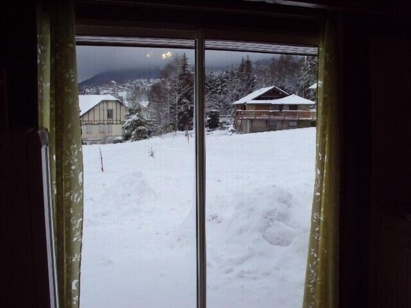 Ett snötäckt landskap med ett hus och ett berg i bakgrunden, sett genom ett fönster med en gardin.