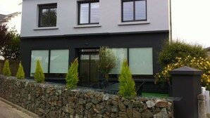 A modern two-story house with a stone wall, a black roof, and a small garden with yellow flowers.