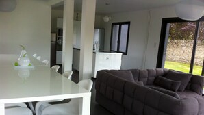 A modern living room with a glass dining table, white chairs, a dark grey sofa, and a kitchen area in the background.