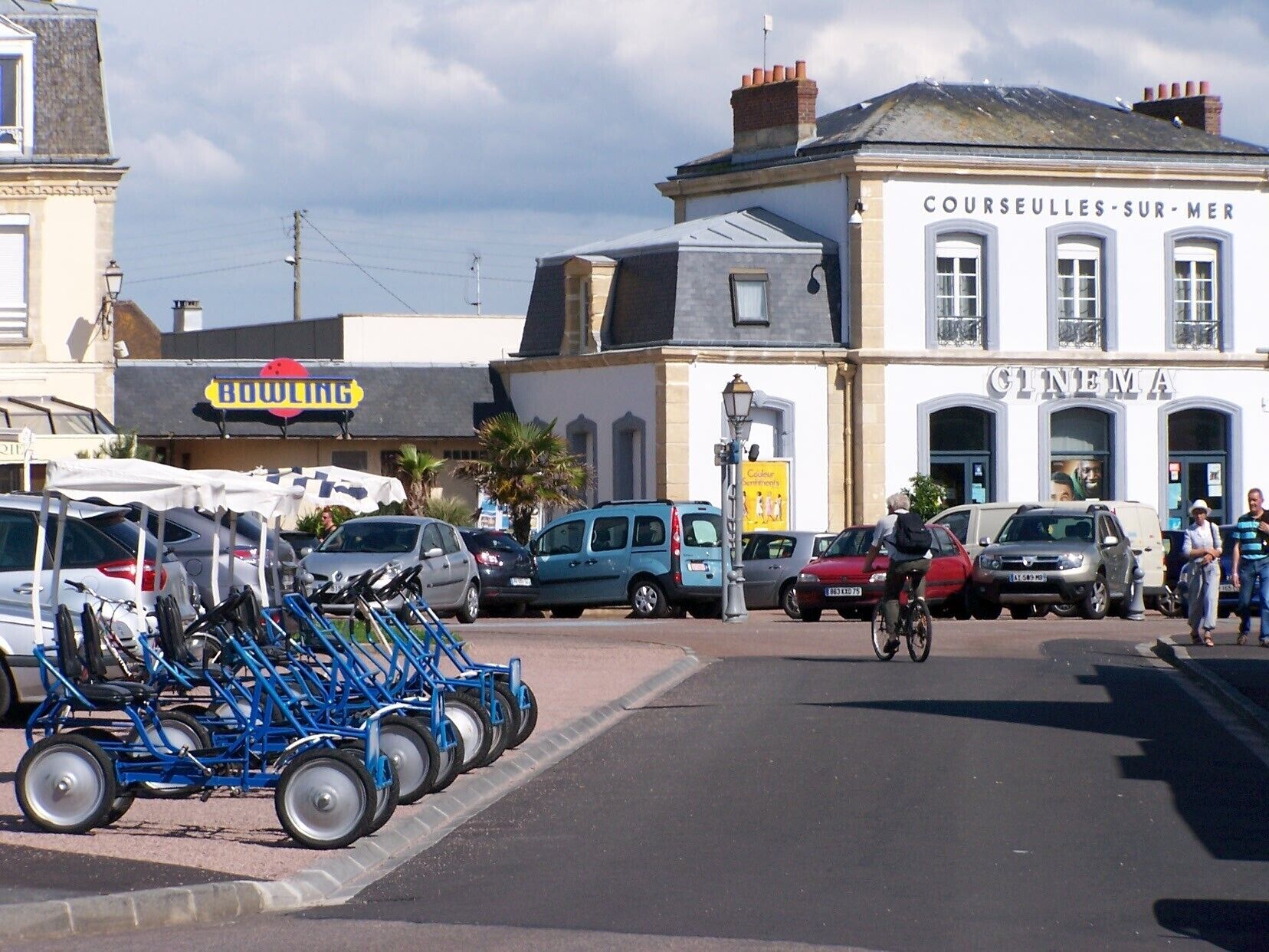 A street scene with parked cars, a bicycle cart, and buildings including a cinema and a bowling alley.