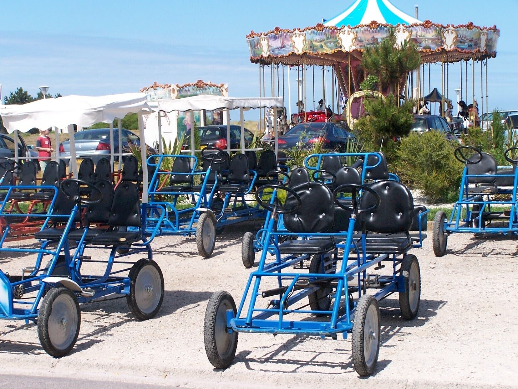 Une rangée de chariots bleus à trois roues, dotés de sièges noirs, est garée sur une surface sablonneuse.