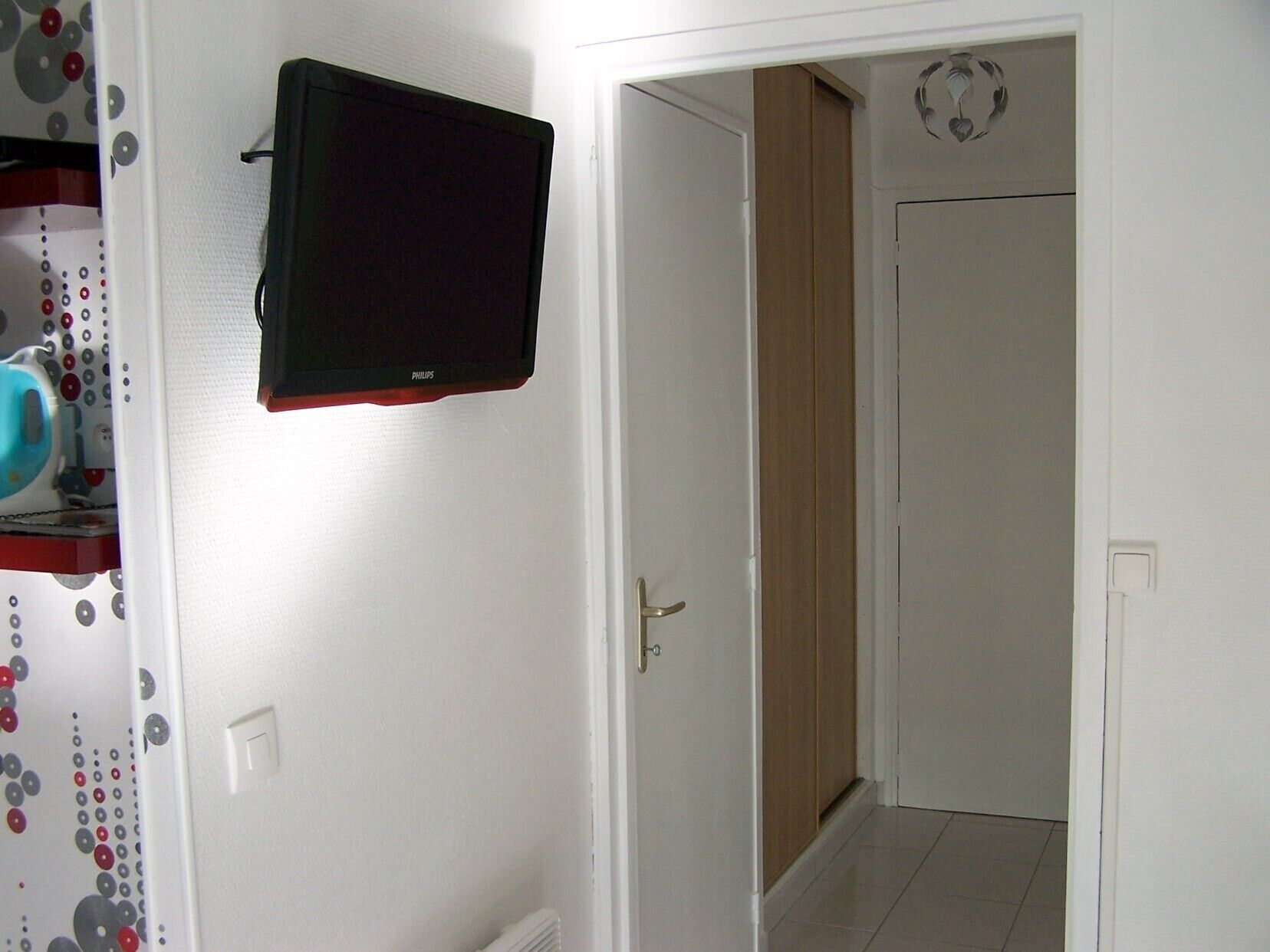 A room with a television mounted on the wall, a door leading to another room, and a patterned curtain on the left.