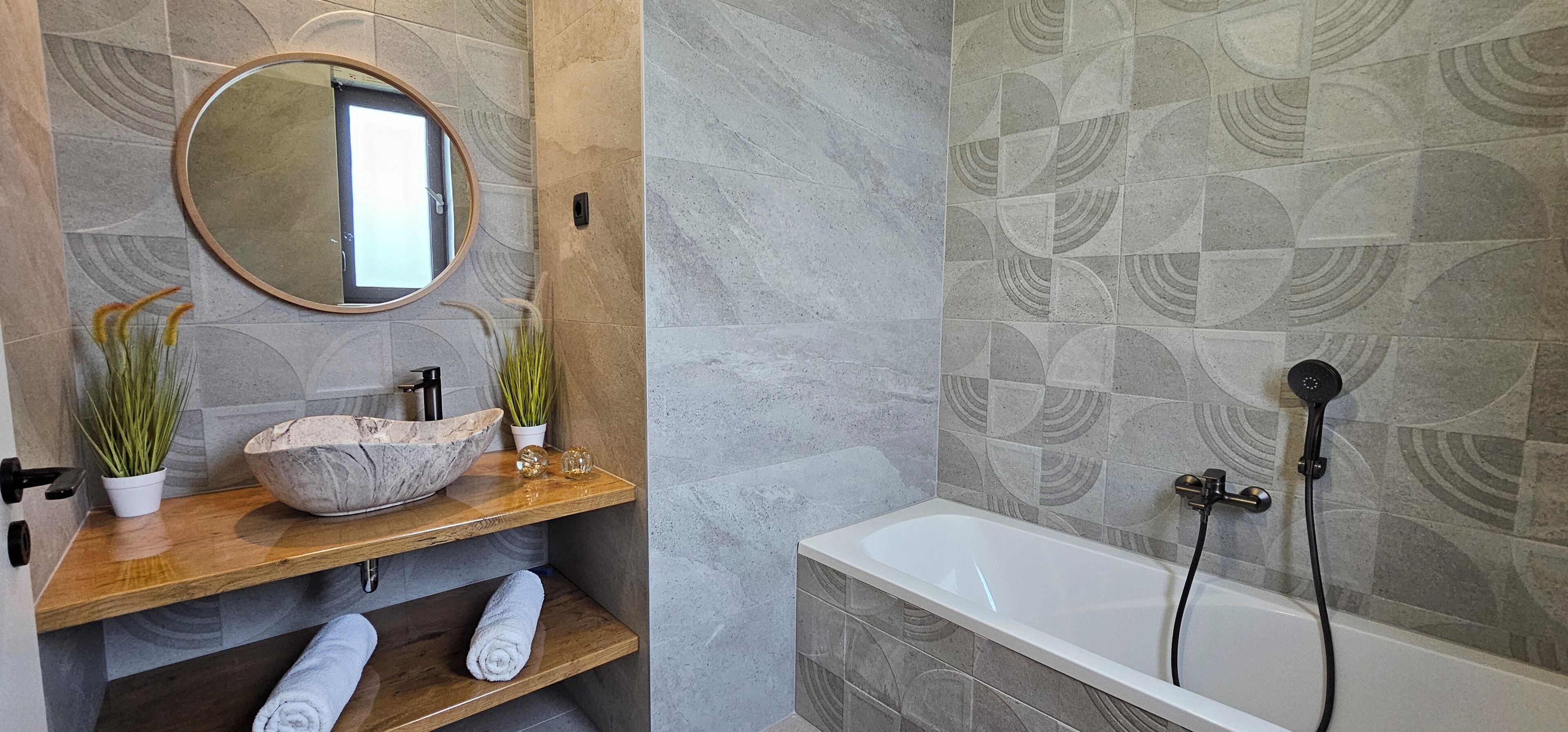 Une salle de bain avec un lavabo en marbre, un meuble-lavabo en bois, deux plantes en pot, un miroir rond et une baignoire.