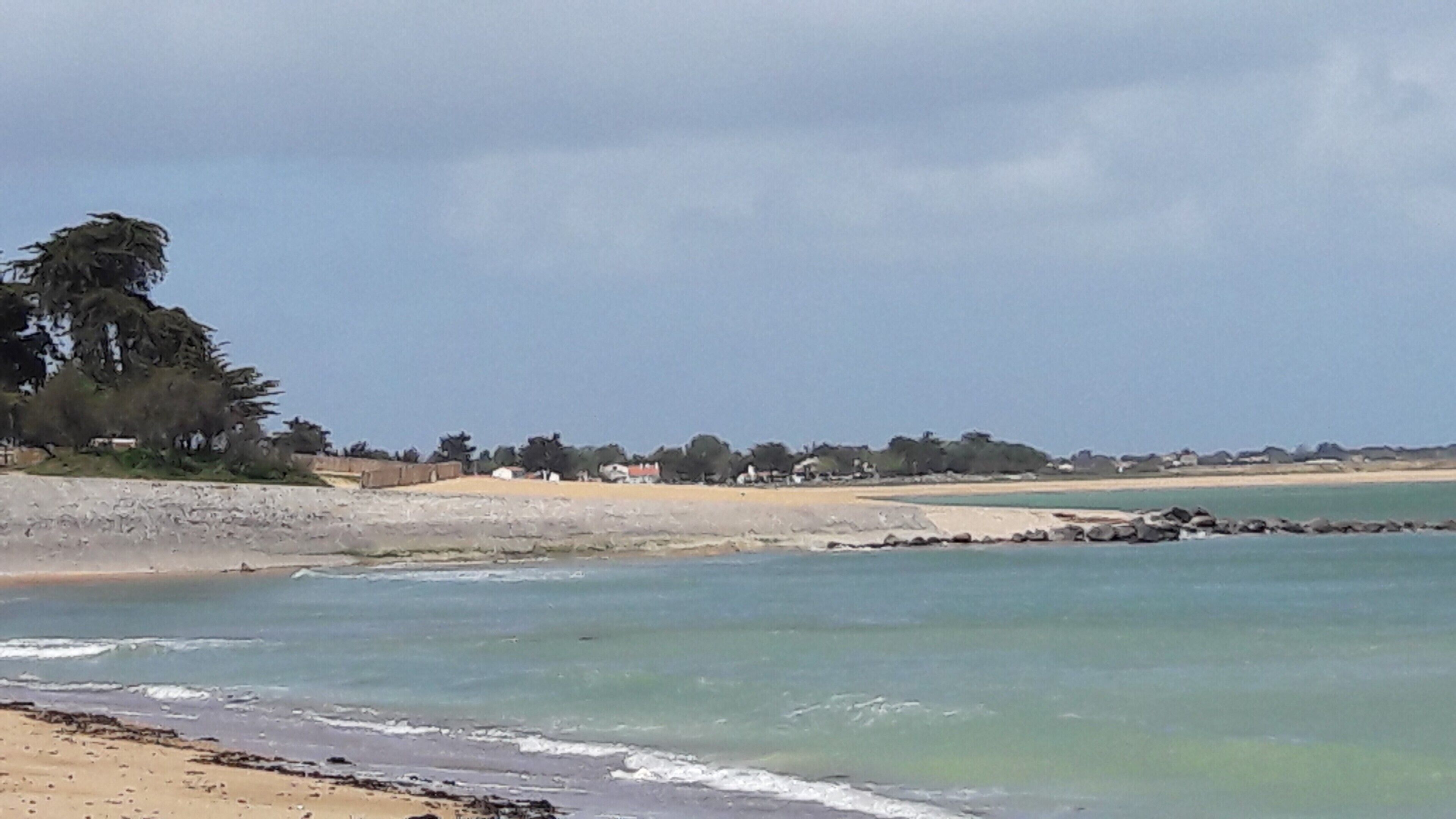 A coastal beach scene with a sandy shore, calm sea, and a row of houses in the background.