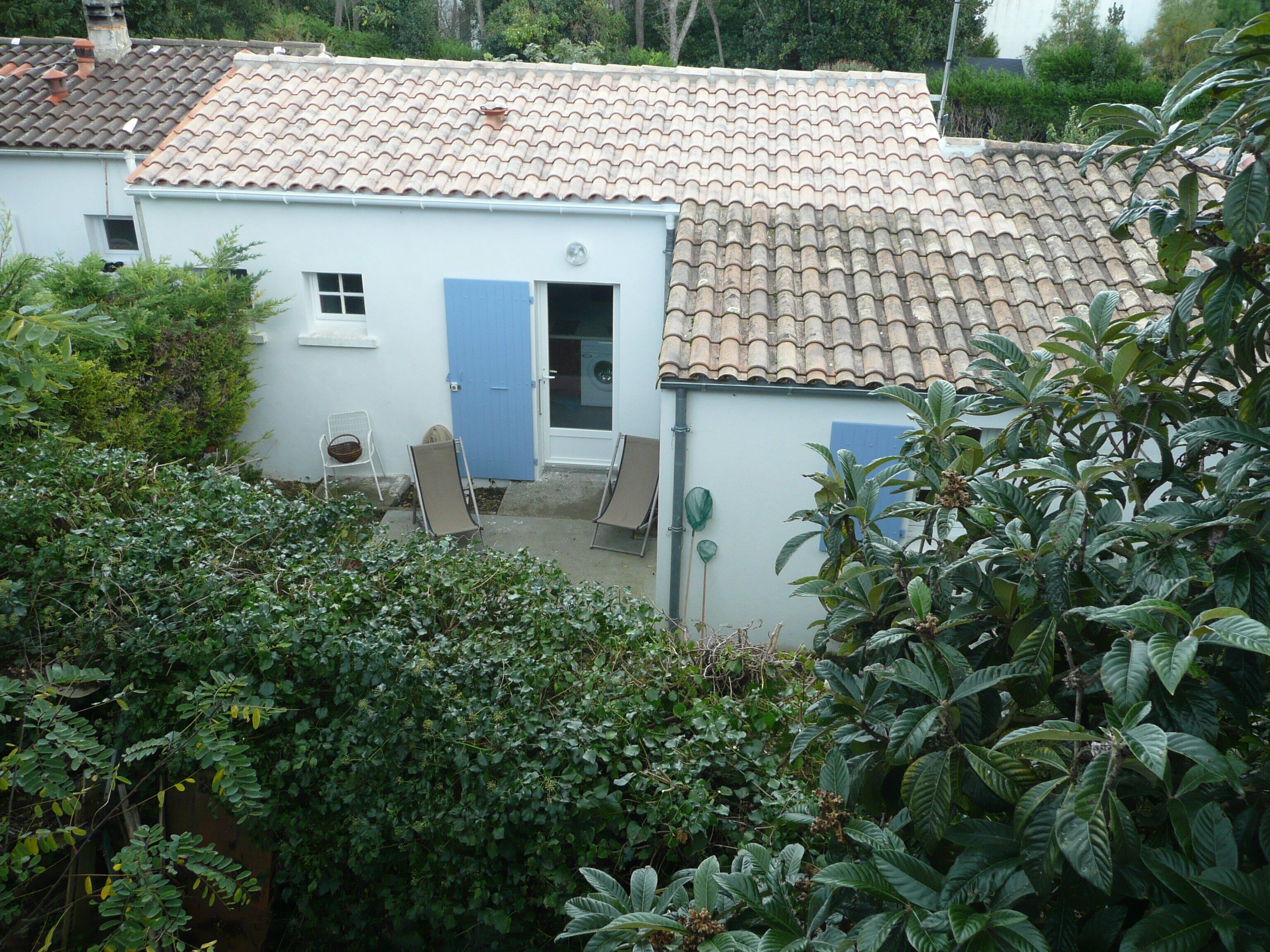 A white building with a tiled roof, a blue door, and a small window. There are two chairs outside and a potted plant nearby.