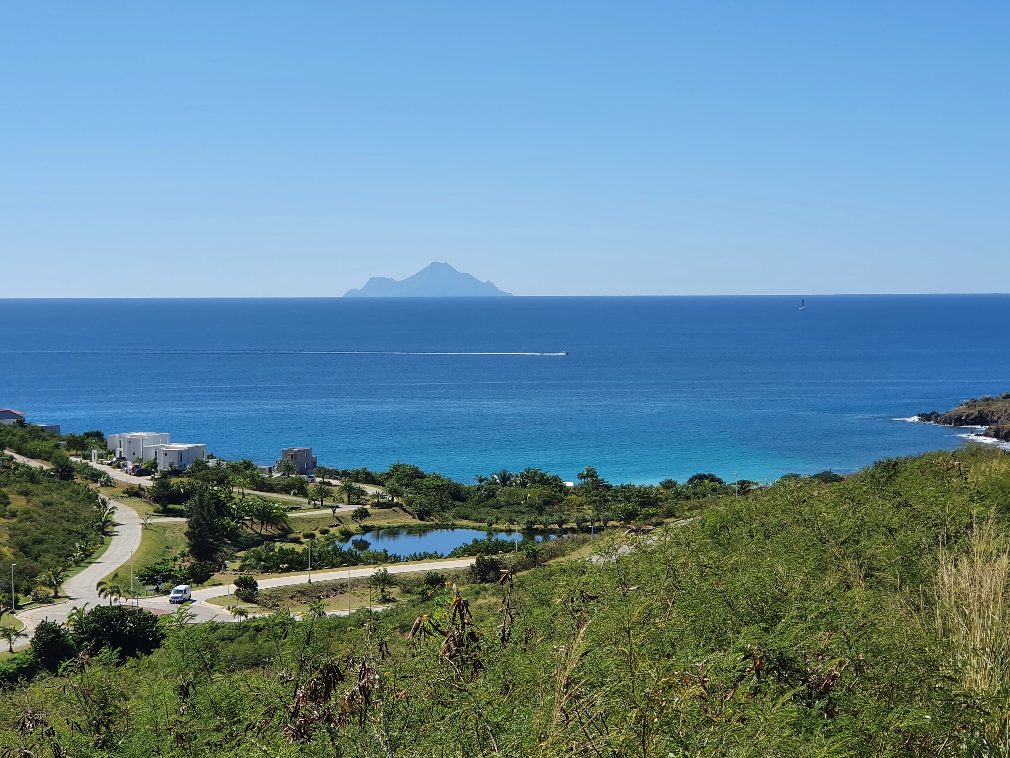 A coastal landscape with a clear view of the ocean, a distant mountain, and a road leading to a building complex.