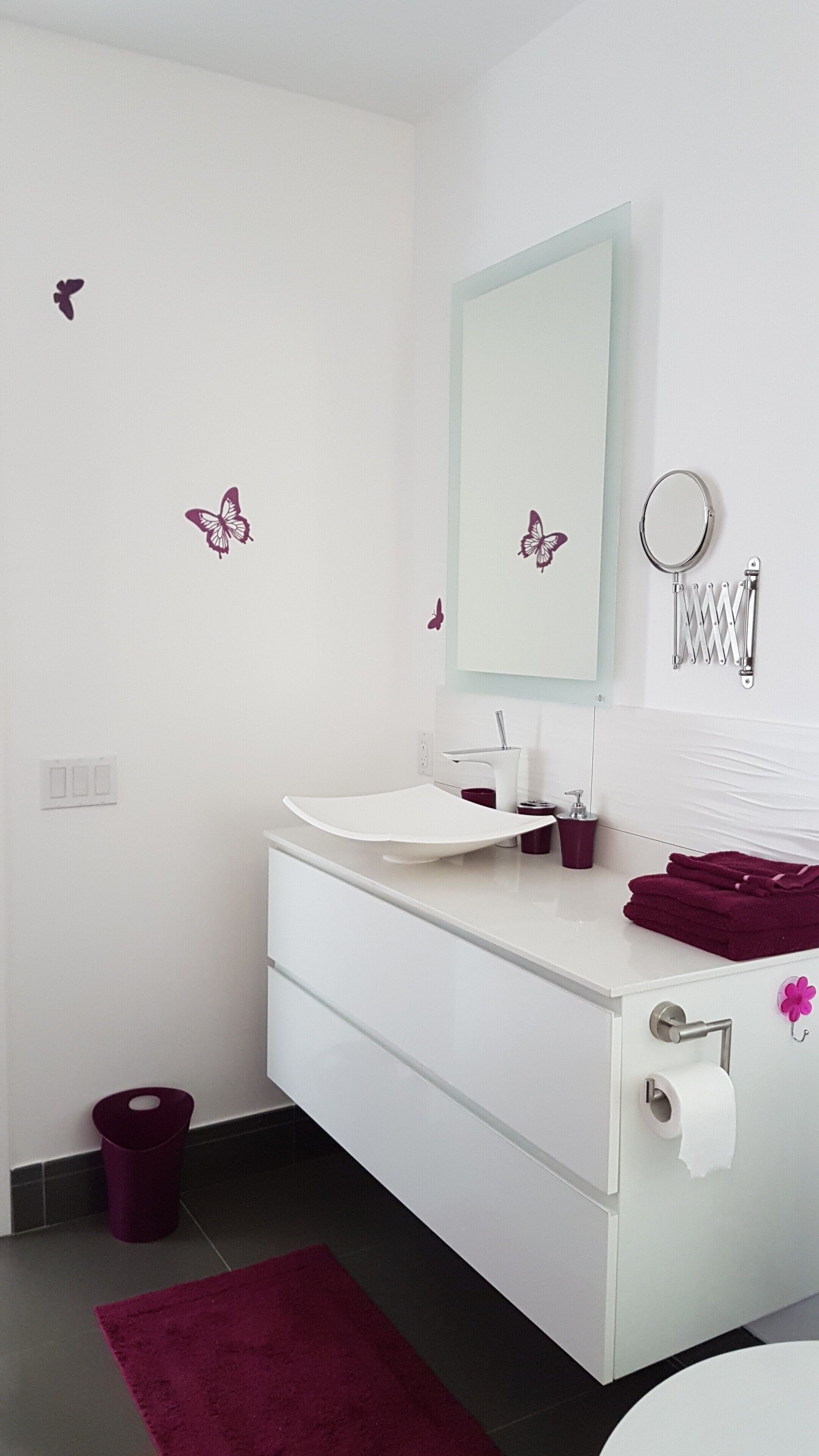 Un bany amb un lavabo blanc, un mirall, un rentapeus i un vàter, i una catifa de color morat.