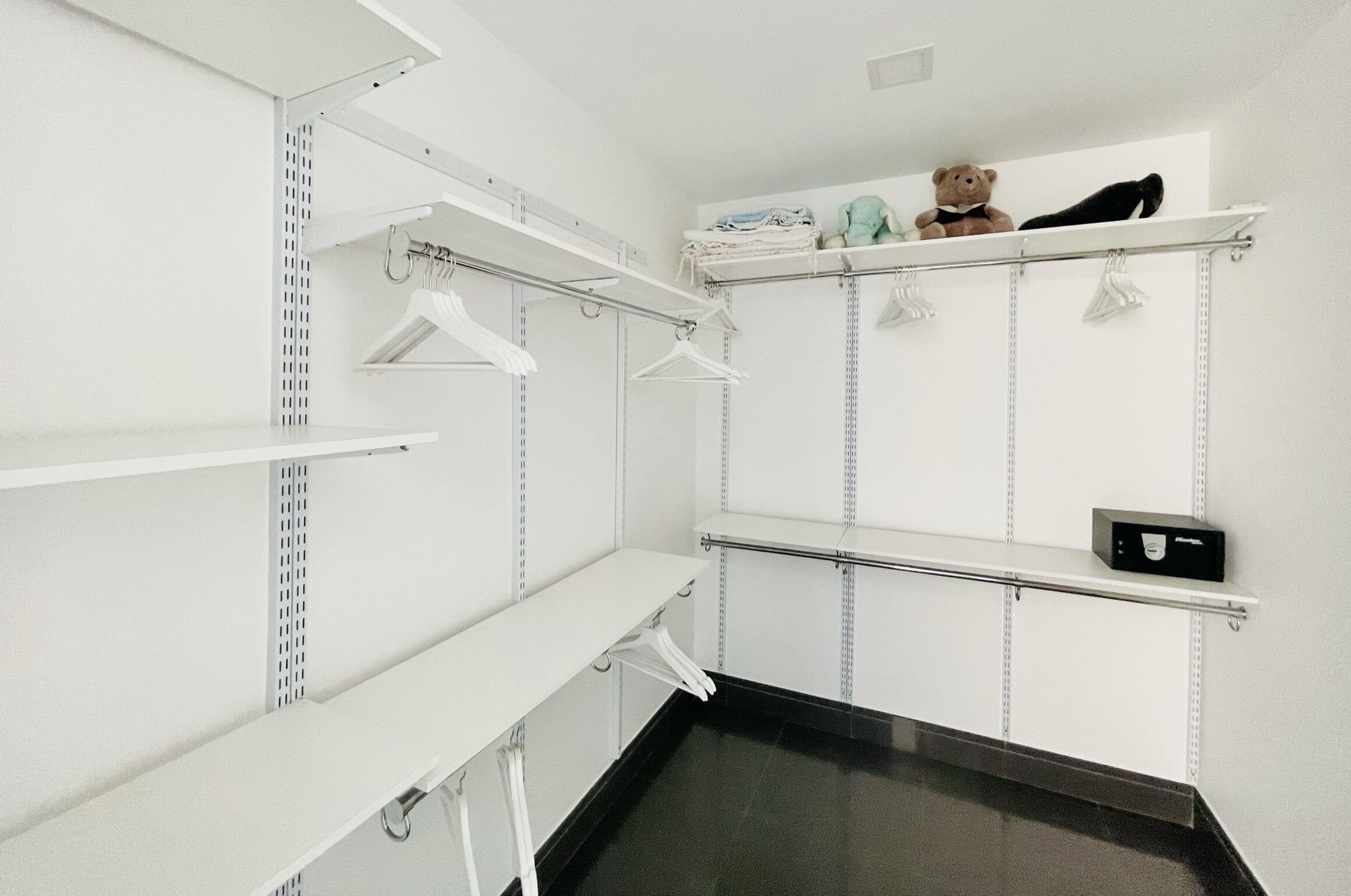 A walk-in closet with white shelving units, a black countertop, and a teddy bear on the top shelf.