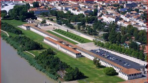 A large, rectangular building with a long, straight facade, adjacent to a river. There are multiple parking spaces visible in front of the building. - Charming house near the beach, detached, not overlooked, WIFI. (Arvert)