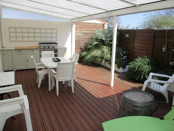 En overdækket terrasse med træterrasse, hvide møbler, grill og plantekasser.