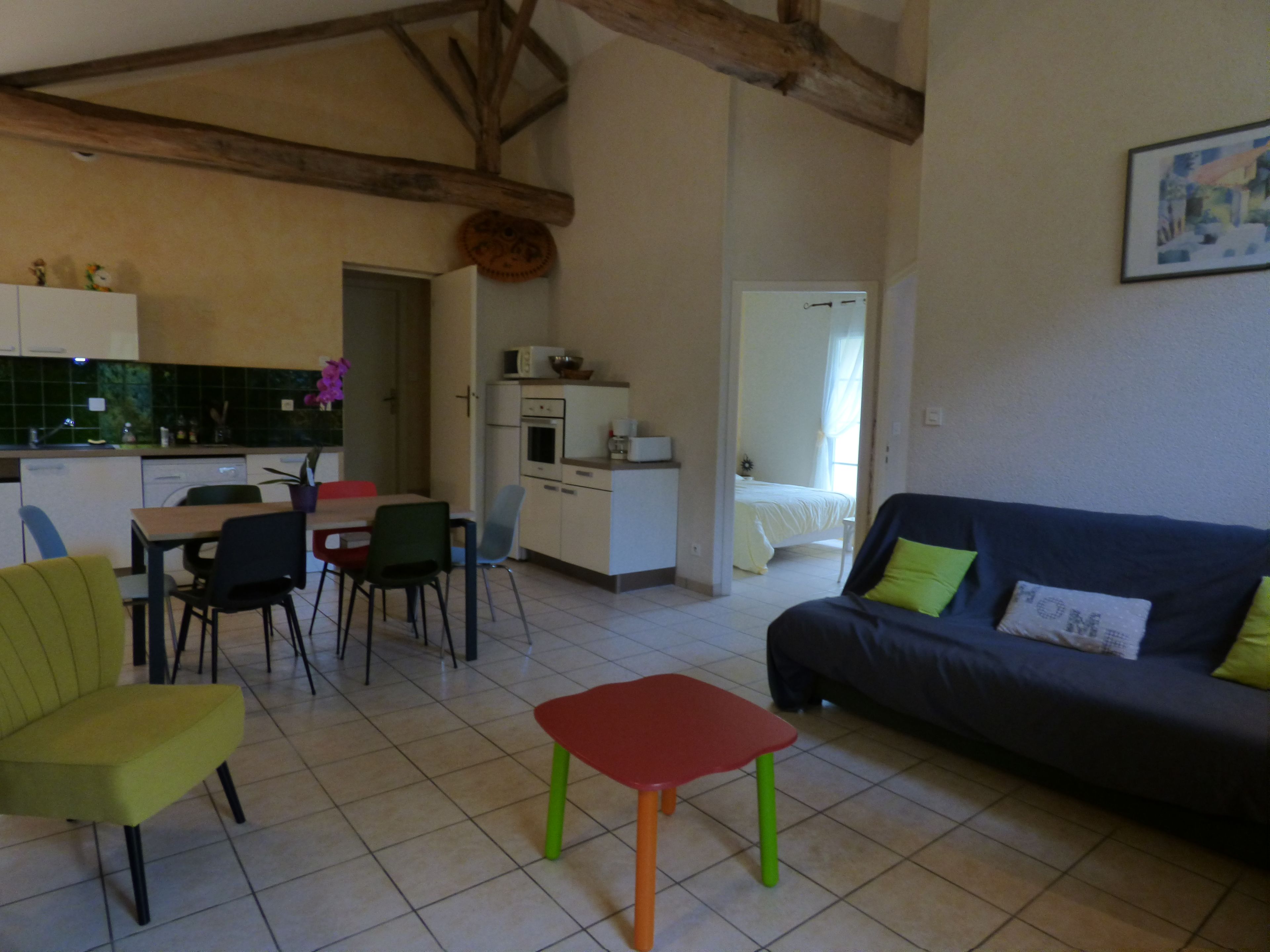 Un salon avec un canapé, des fauteuils, une petite table, un coin cuisine avec des placards et des appareils électroménagers, et une chambre à coucher avec un lit et une fenêtre.