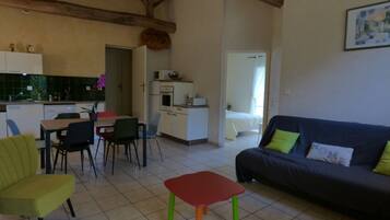 Un salon avec un canapé, des fauteuils, une petite table, un coin cuisine avec des placards et des appareils électroménagers, et une chambre à coucher avec un lit et une fenêtre.
