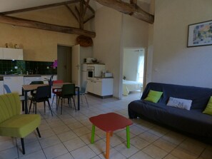 Una sala de estar con sofá, sillas, una mesita, un área de cocina con alacenas y electrodomésticos, y un dormitorio con cama y ventana.