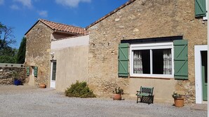 Un rústico edificio de piedra con tejado de baldosas, una ventana con contraventanas verdes y una banca afuera.