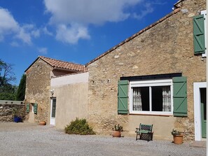 Un bâtiment rustique en pierre, avec un toit en tuiles, une fenêtre ornée de volets verts et un banc à l’extérieur.