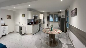Cocina compacta con armarios blancos, una mesa de comedor con sillas transparentes y un ventanal con cortinas.