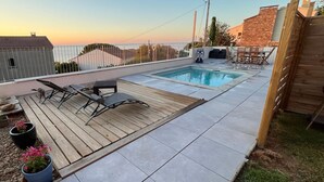 Un área de piscina en la azotea con terraza de madera, sillones de descanso y vistas al atardecer.