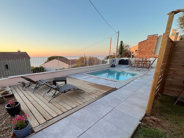 A rooftop pool area with a wooden deck, lounge chairs, and a view of the sunset.