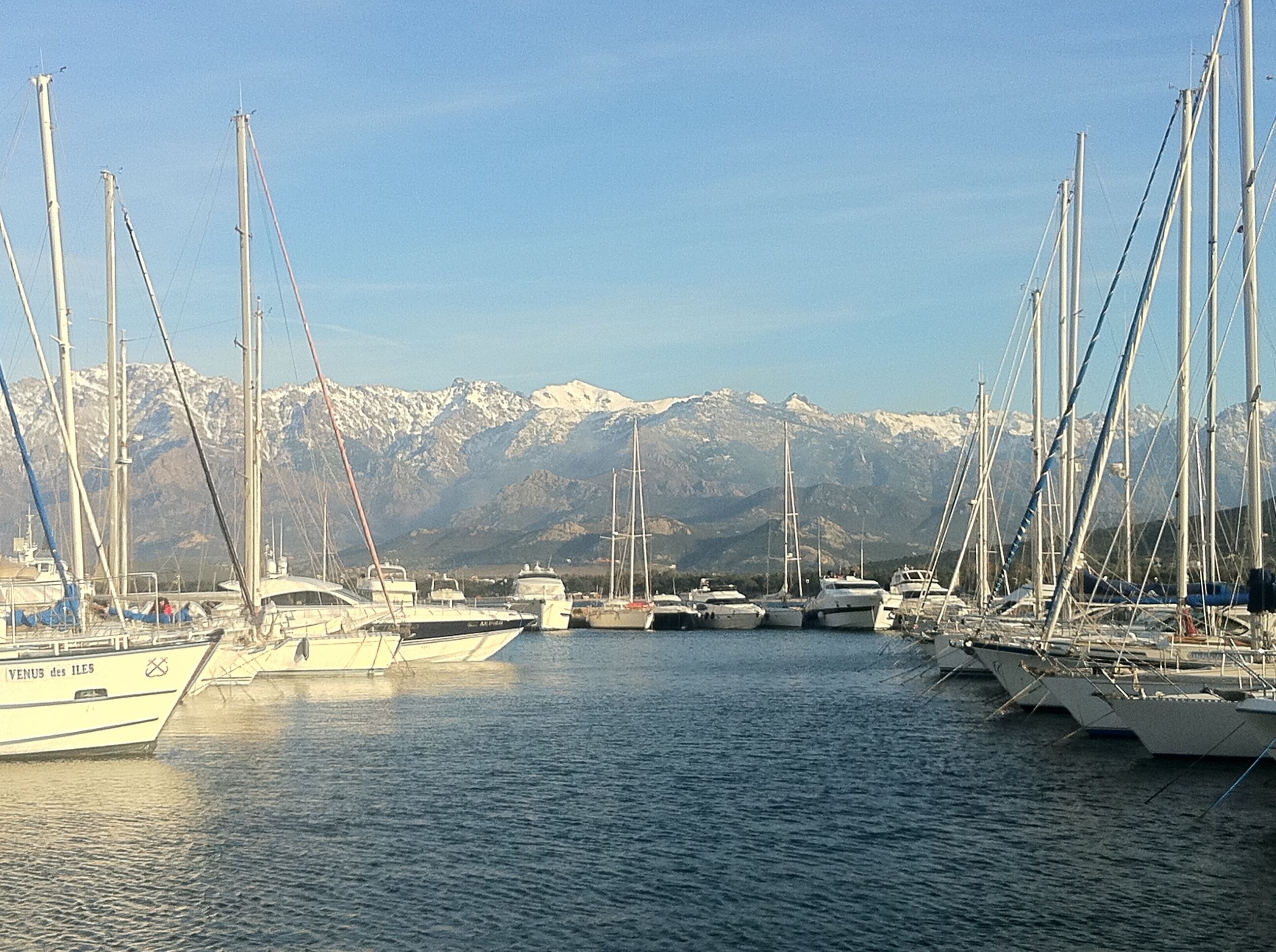 Ein Yachthafen mit mehreren Segelbooten, eine Bergkulisse im Hintergrund und strahlend blauer Himmel.
