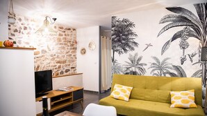 A living room with a stone wall, a green sofa with patterned cushions, a wooden TV stand, and a wall mural of tropical plants. - Superb apartment (3pers) air-conditioned, quiet, ideal location in town center (Nîmes)
