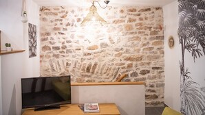 A room with a stone wall, a flat-screen TV, a wooden table, and a hanging light fixture. - Superb apartment (3pers) air-conditioned, quiet, ideal location in town center (Nîmes)