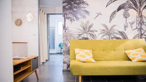 A living room with a green sofa, two yellow patterned pillows, a wooden side table, and a wall with a tropical wallpaper. - Superb apartment (3pers) air-conditioned, quiet, ideal location in town center (Nîmes)