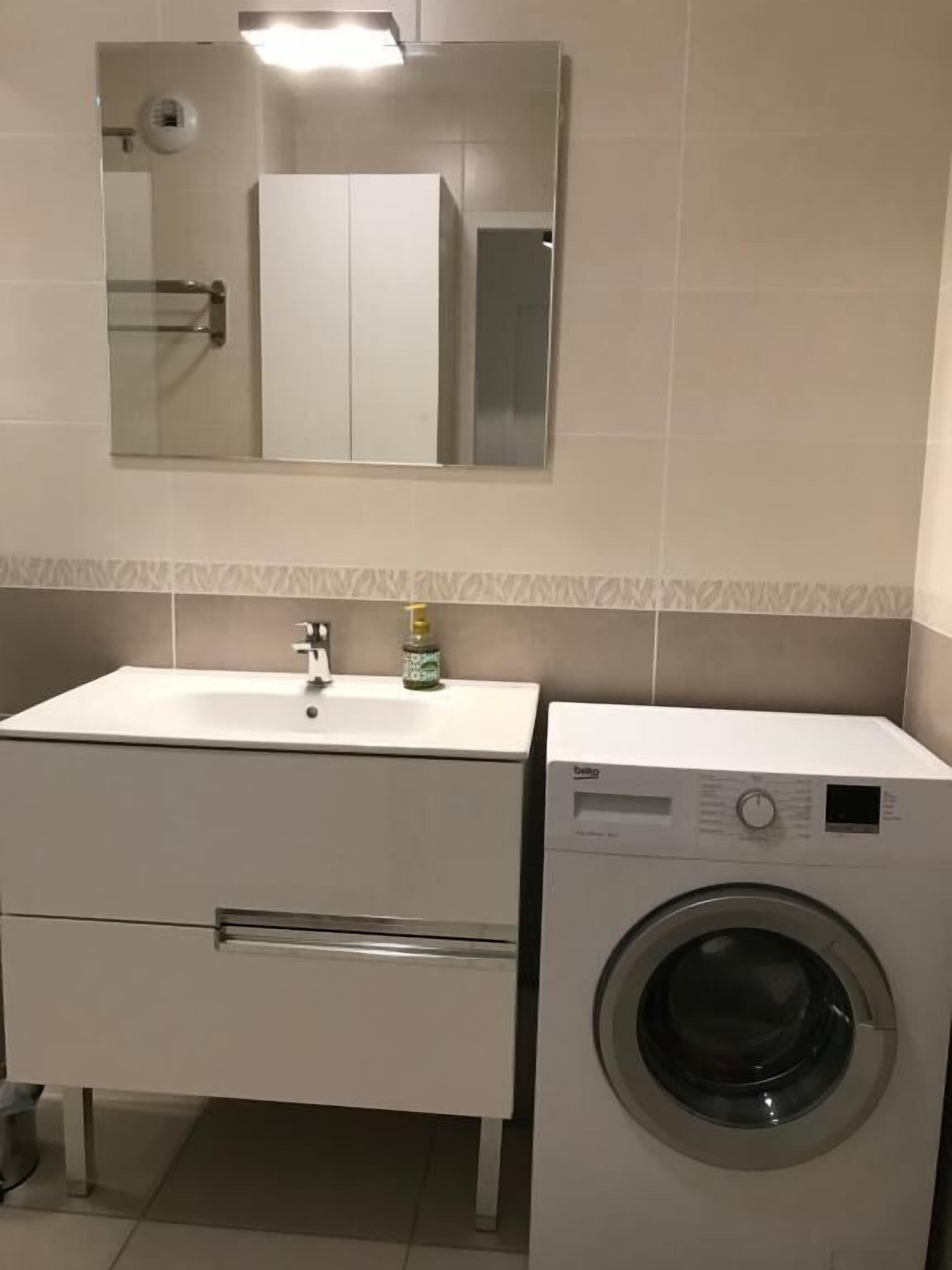 Ein kleines Badezimmer mit einem weißen Waschbecken, einem Spiegel darüber und einer Waschmaschine mit Frontlader.