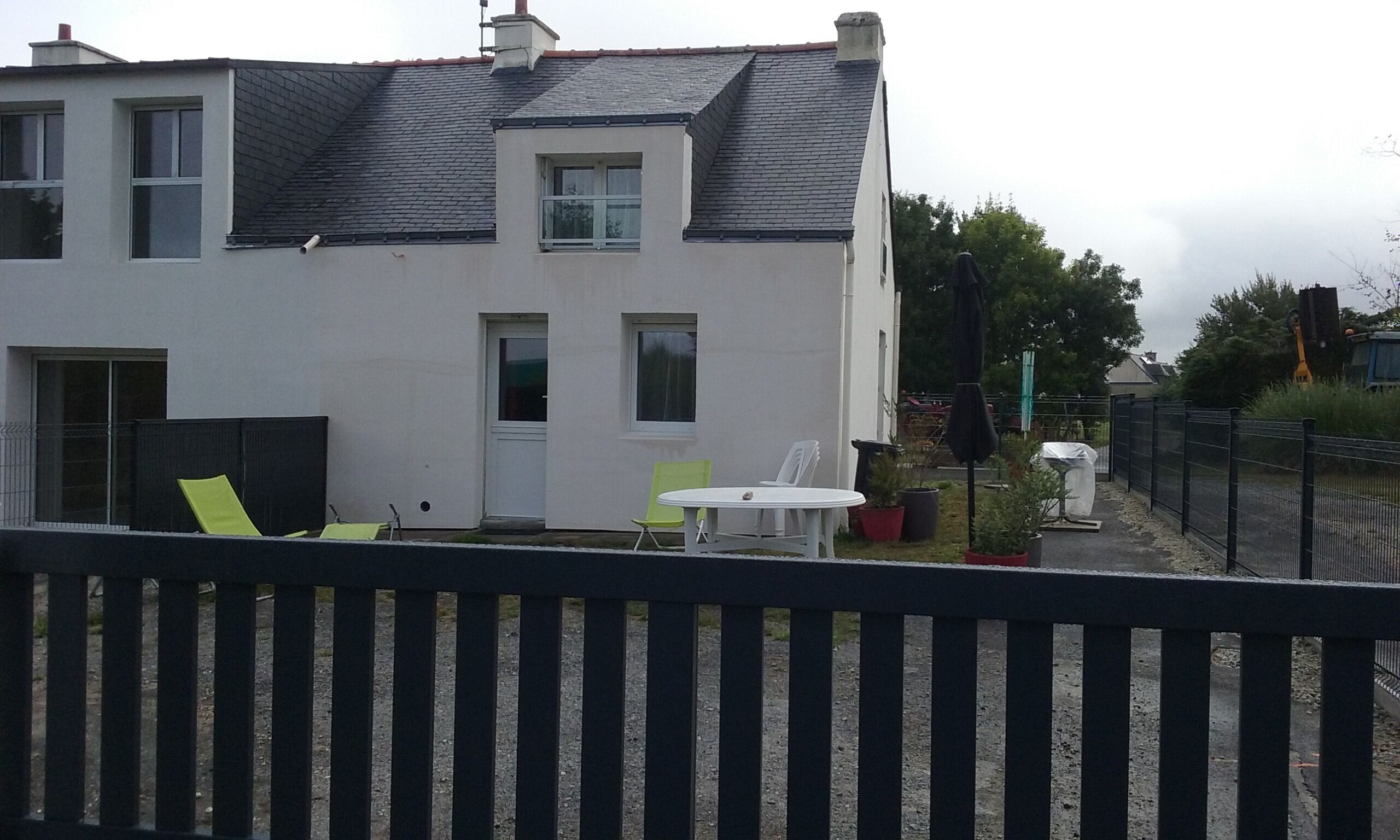 A two-story house with a white exterior, a dark roof, and a fenced backyard. There are two windows on the ground floor and one on the upper floor.