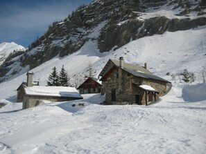 A stone cabin with a sloped roof, surrounded by snow-covered ground and a mountainous backdrop. - Guillestre ground-floor rental for 2 to 4 people, beautiful south-facing view. (Guillestre)