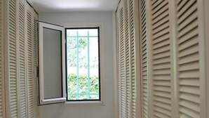 A narrow hallway with white tiled flooring, a window with a metal grid, and white louvered closets on both sides.