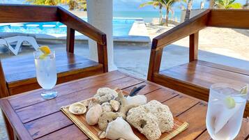 A wooden table with a tray of seashells and a knife, two glasses of water with lemon slices, and a view of the beach and ocean.